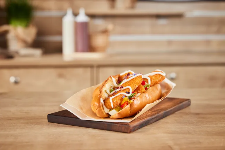 Ein Hotdog mit knusprigen Orangenstückchen, garniert mit Mayonnaise und Gemüse, auf einem Holzbrett vor einem verschwommenen Küchenhintergrund.