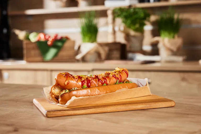 Nahaufnahme eines Hotdogs mit Ketchup und knusprigen Zwiebeln auf einem Holztablett, im Hintergrund sind verschwommene Küchenregale zu sehen.