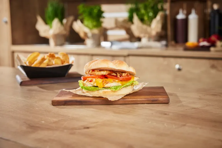 Ein Burger mit Salat, Tomate und Käse auf einem Holzbrett, im Hintergrund dazu Kartoffelecken.