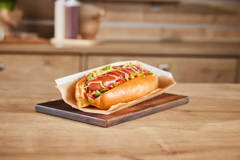 Ein Gourmet-Hotdog mit Senf, Zwiebeln und Paprika als Belag, serviert in einer Papierschale auf einem Holzbrett, vor dem Hintergrund einer Küche.