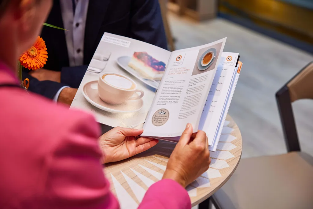 Eine Person in rosa Kleidung liest an einem Tisch, auf dem eine leuchtend orangefarbene Gerbera steht, eine Speisekarte mit einem Cappuccino-Foto und einem Text über die Zubereitung von Kaffee.