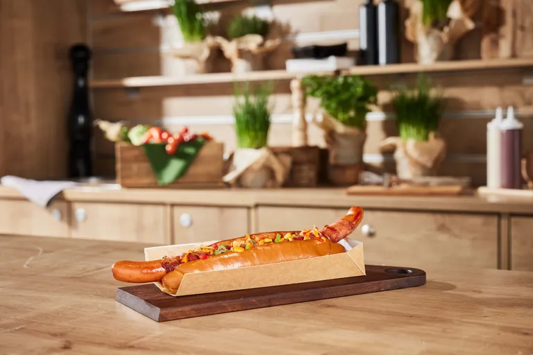 Ein Gourmet-Hotdog mit Belag, serviert in einer Papierschale auf einem Holzbrett in einer gemütlichen Küchenatmosphäre.