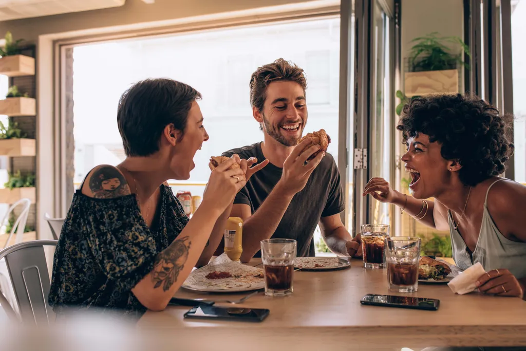Drei Freunde lachen gemeinsam, während sie an einem gemütlichen Restauranttisch, umgeben von Tageslicht und Pflanzen, Burger und Getränke genießen.