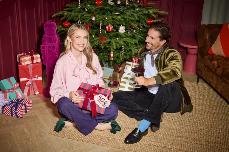 Eine Frau und ein Mann sitzen, festlich gekleidet, vor einem Weihnachtsbaum am Boden, umringt von Weihnachtspaketen und Dekoration. Er hält ein Weinglas und sie ein Geschenk in den Händen.