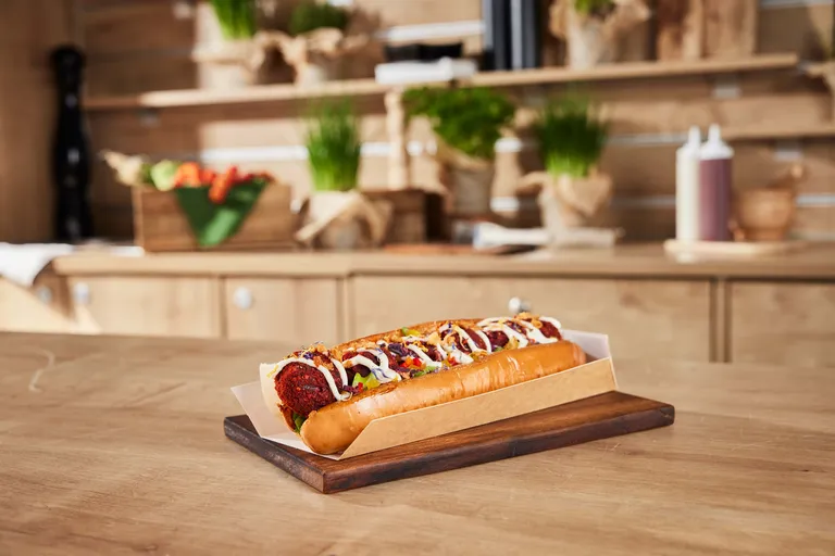 Ein reichhaltig belegter veganer Hotdog mit Senf und Zwiebeln auf einem Holzbrett in einer rustikalen Küchenumgebung.