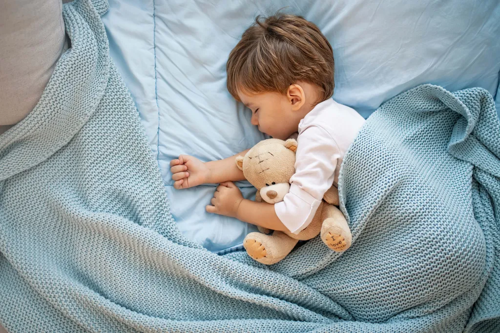 Kleines Kind schläft zugedeckt im Bett und hält einen Teddybären im Arm