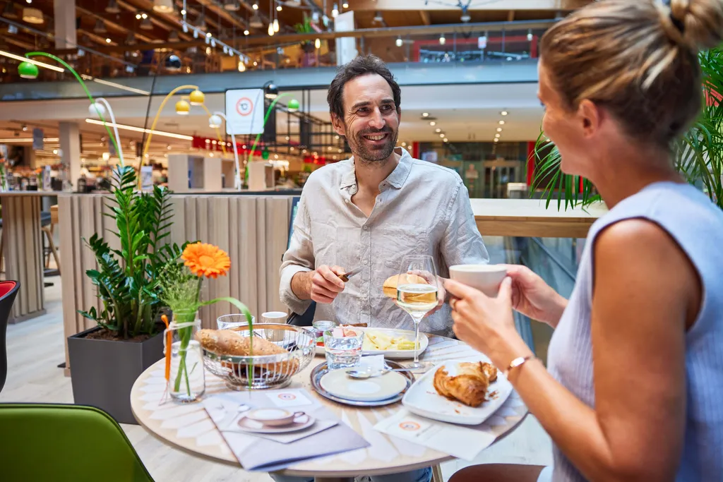 Ein Mann und eine Frau genießen in einem Café ihr Frühstück, zu dem Croissants und Wein gehören. Das Ambiente ist modern, mit heller Einrichtung und Pflanzen.