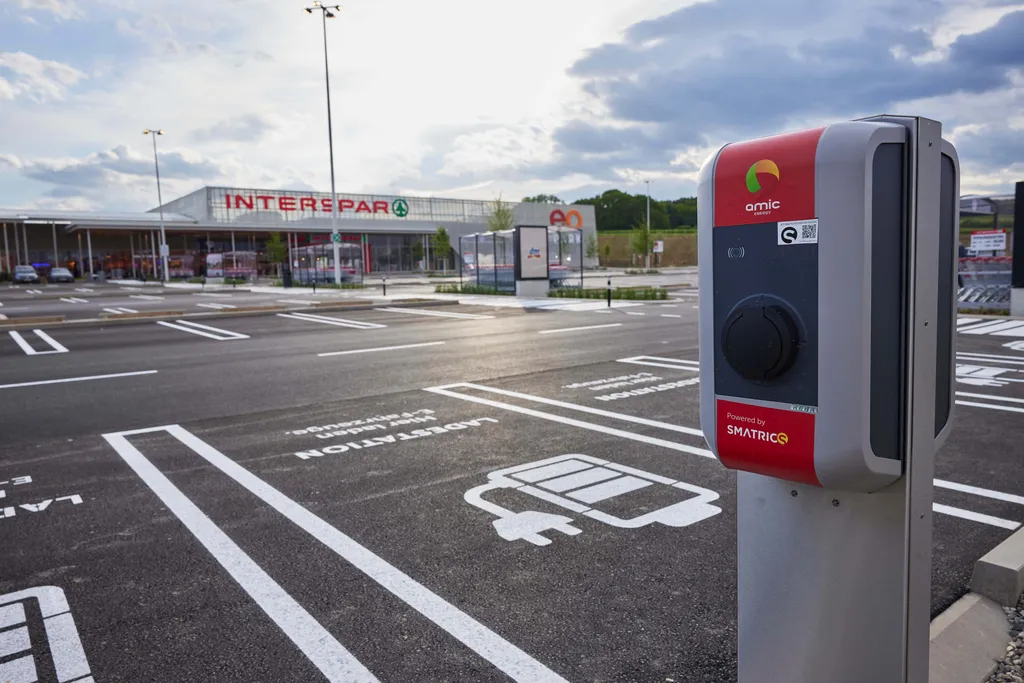 Eine Ladestation für Elektrofahrzeuge auf einem Parkplatz vor einem Interspar-Supermarkt, bei klarem Himmel und freien Parkplätzen.
