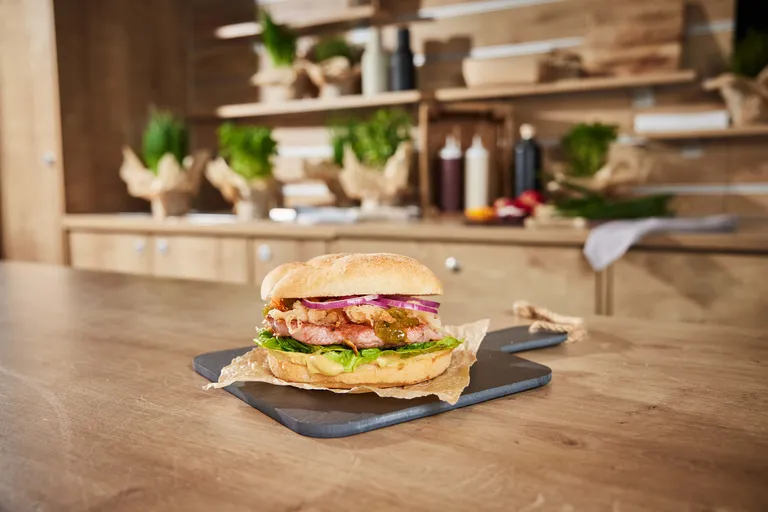 Ein leckerer Burger mit Speck, Zwiebeln und Salat auf einem Ciabatta-Brötchen, serviert auf einem Schieferbrett in einer rustikalen Küchenumgebung.