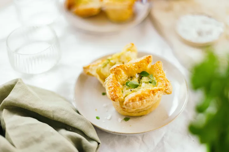 Mini-Törtchen mit goldbrauner Kruste und grüner Garnitur auf einem weißen Teller, umgeben von einer grünen Serviette und einem Glas.