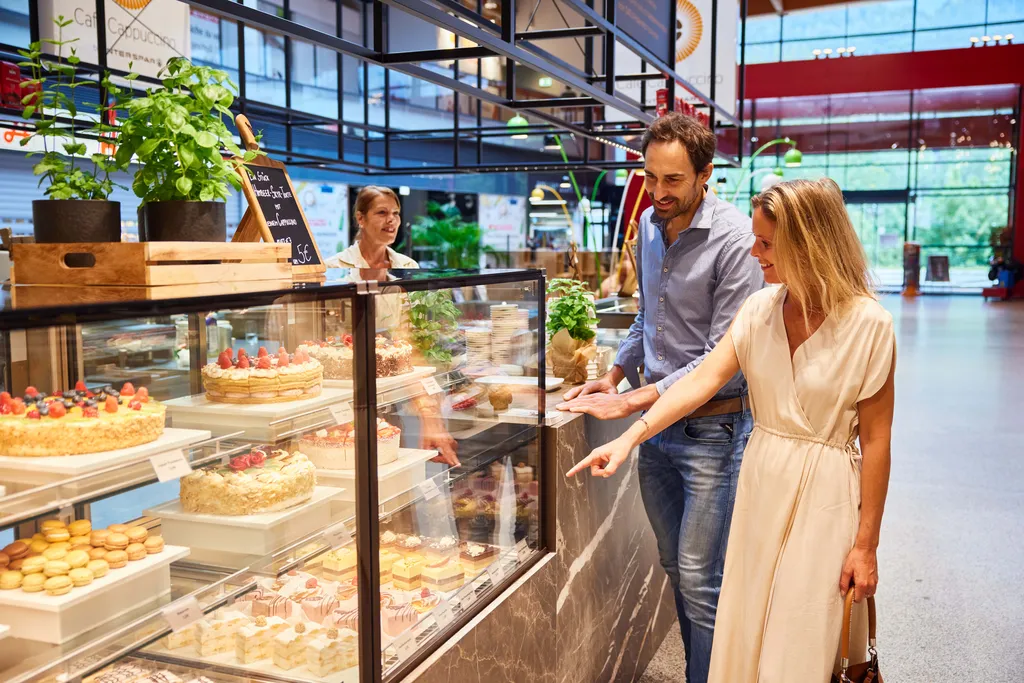 Ein Paar bewundert Kuchen und Gebäck in der Auslage einer Bäckerei, während der Ladenbesitzer lächelnd danebensteht.