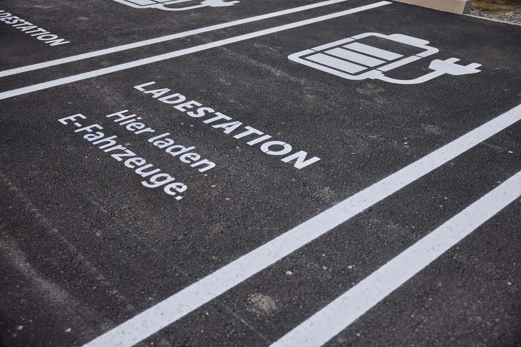 Parkplatz mit Markierungen für das Laden von Elektrofahrzeugen, beschriftet mit „Ladestation“ und „Hier laden E-Fahrzeuge“ sowie einem Ladesymbol.