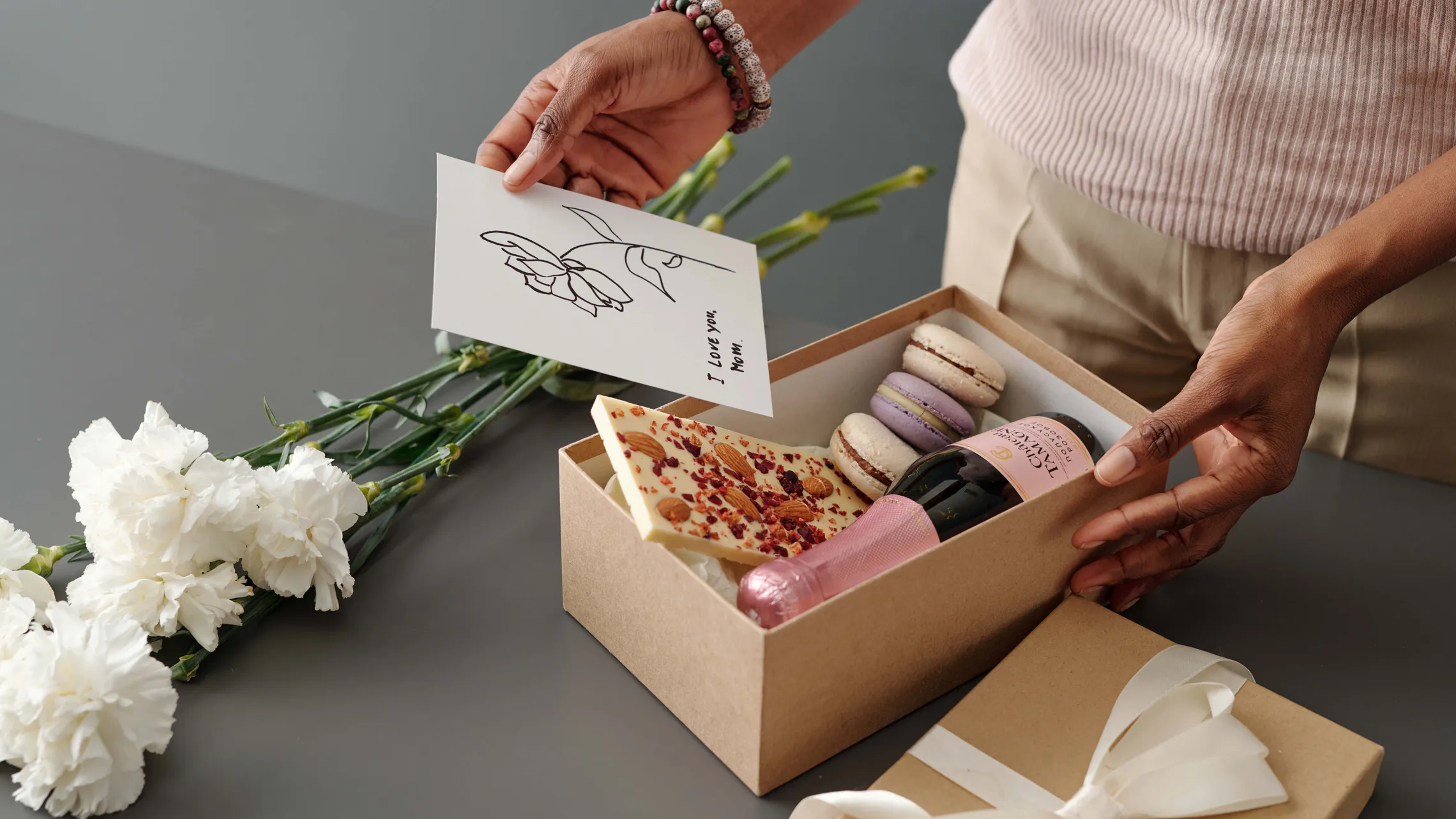 Eine Frau verpackt ein Muttertagsgeschenk bestehend aus einer Box mit Schokolade und Macarons, Sekt und Nelken, darauf legt sie eine Karte