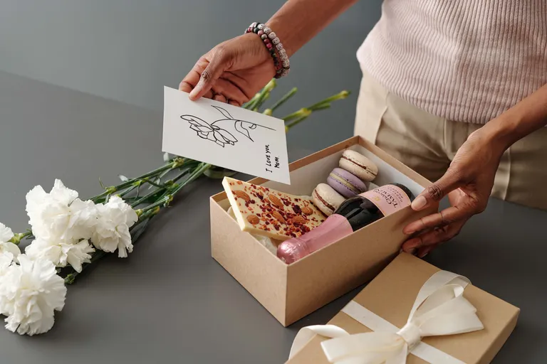 Eine Frau verpackt ein Muttertagsgeschenk bestehend aus einer Box mit Schokolade und Macarons, Sekt und Nelken, darauf legt sie eine Karte