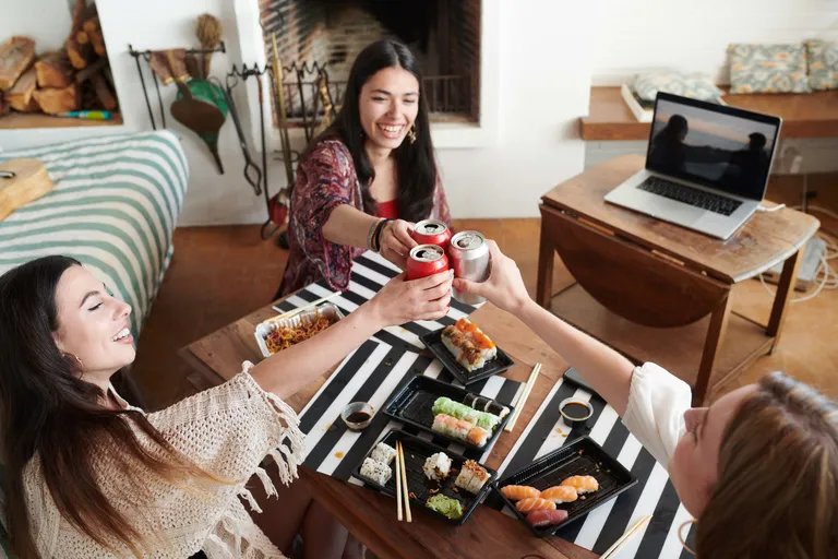 Drei Freunde stoßen bei einem Sushi-Essen zu Hause mit Getränkedosen an, während im Hintergrund auf einem Laptop ein Video läuft.