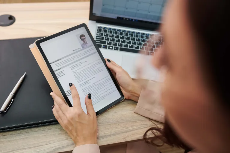 Person liest einen Lebenslauf auf einem Tablet am Schreibtisch mit Laptop