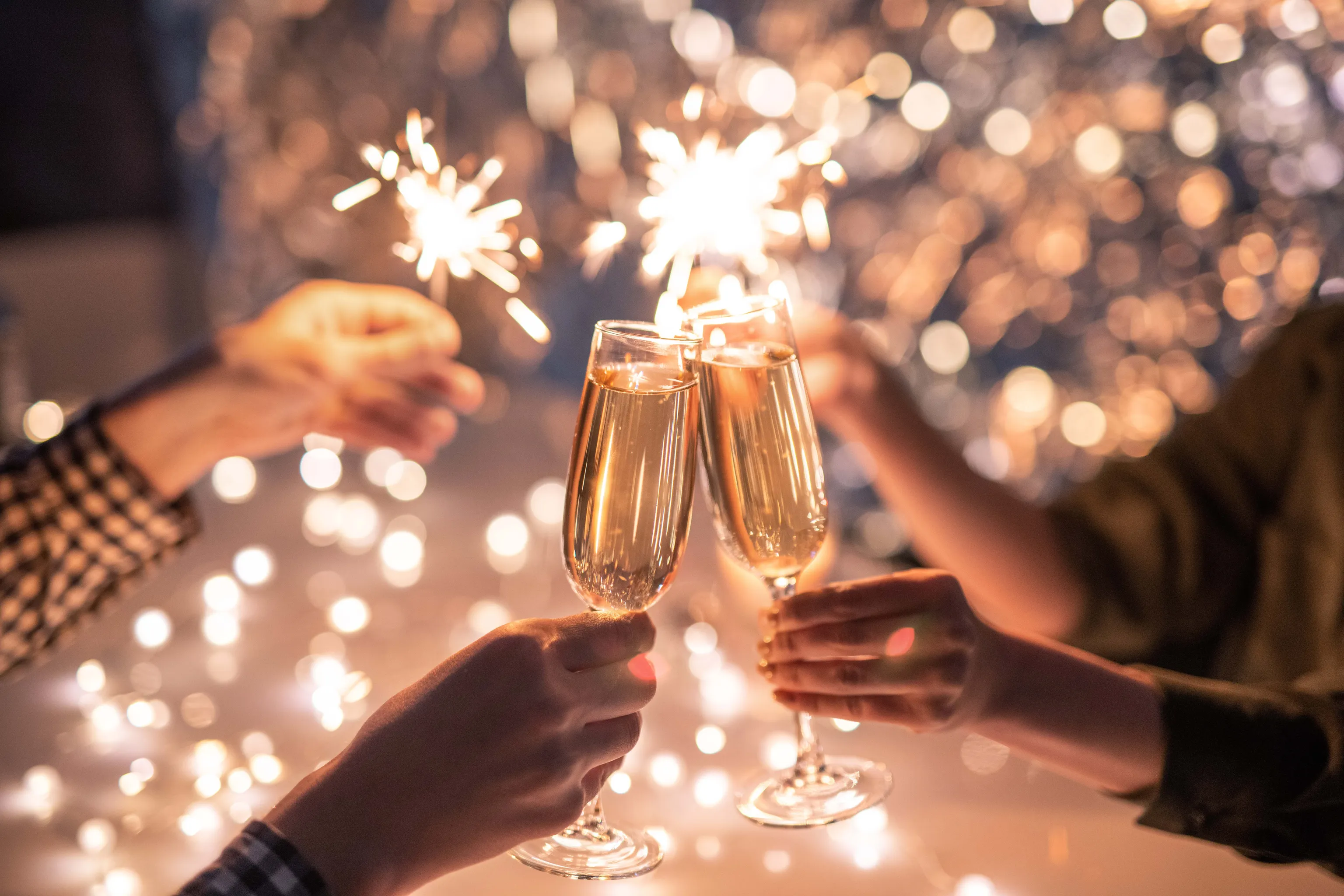 Hände, die Wunderkerzen halten und Champagnergläser anstoßen, vor einem funkelnden, festlichen Hintergrund.