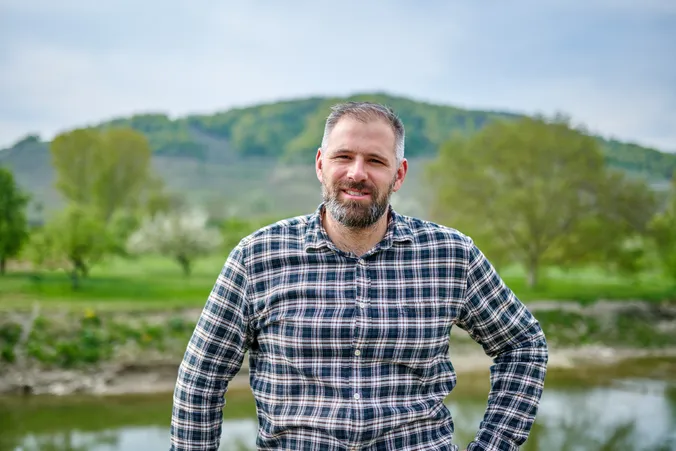 Ein bärtiger Mann in einem karierten Hemd steht im Freien in der Nähe eines Flusses, im Hintergrund sind grüne Bäume und Hügel zu sehen.