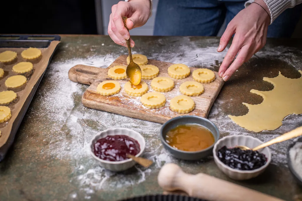 Eine Person verteilt mit einem Löffel Marmelade auf ausgestochenen Mürbteigkeksen. Daneben stehen Schälchen mit verschiedenen Marmeladen.