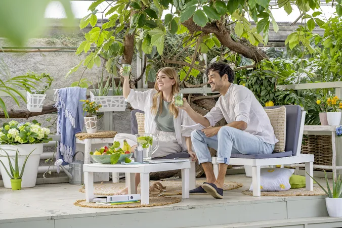 Eine Frau und ein Mann sitzen gemütlich auf Loungemöbeln im Garten und halten Wassergläser in die Luft um jemandem zu zuprosten.