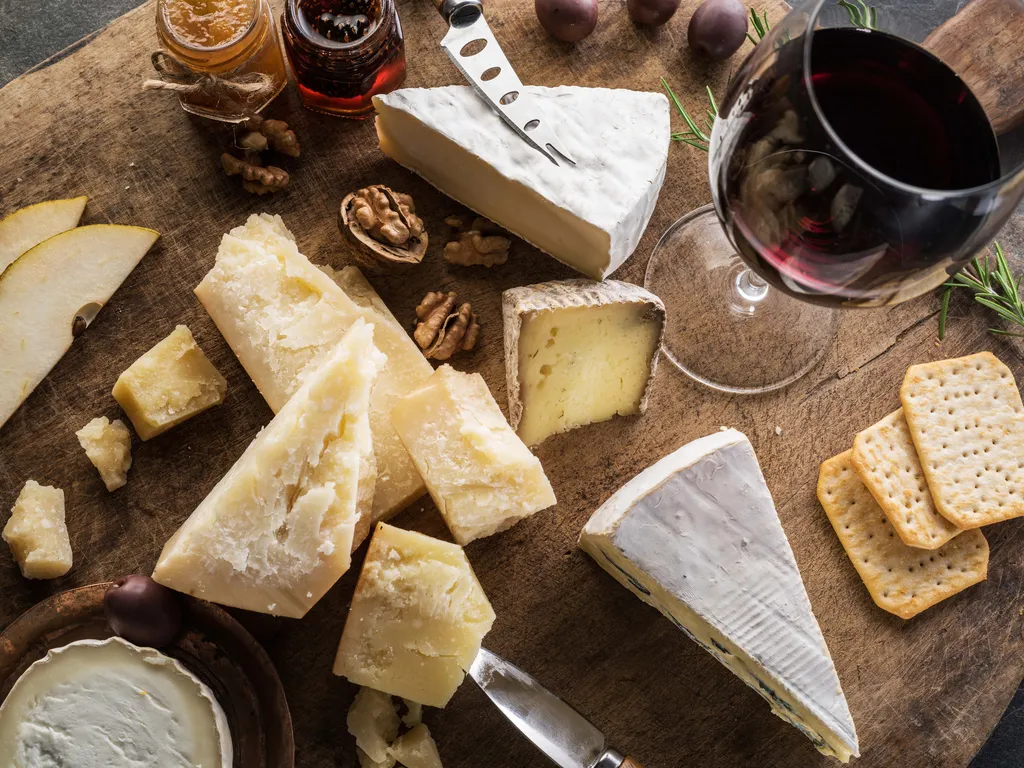 Verschiedene Käsesorten, Walnüsse und Oliven auf einem Holzbrett neben einem Glas Rotwein – eine rustikale Snack-Kombination.