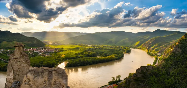 Malerischer Blick auf einen Fluss, der sich durch üppig grüne Hügel und ein Dorf schlängelt, während das Sonnenlicht durch dramatische Wolken am Himmel scheint.