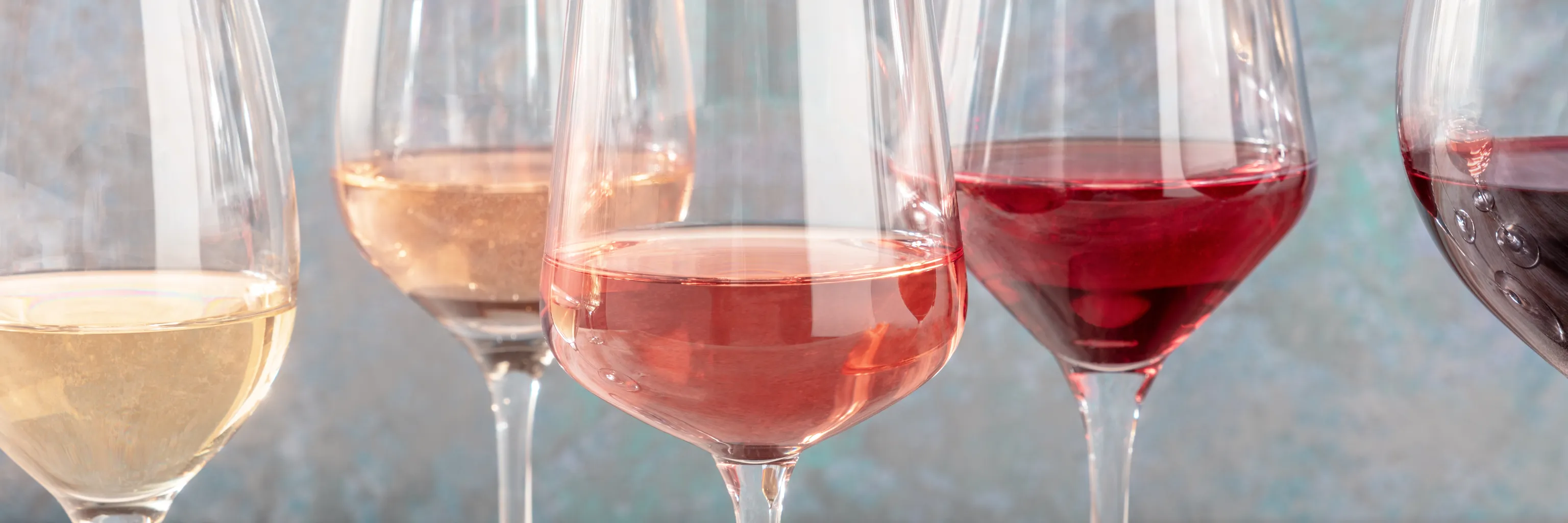 Nahaufnahme von fünf Weingläsern, die mit Wein in verschiedenen Farben – Weißwein, Rosé und Rotwein – gefüllt sind, vor einem unscharfen Hintergrund.