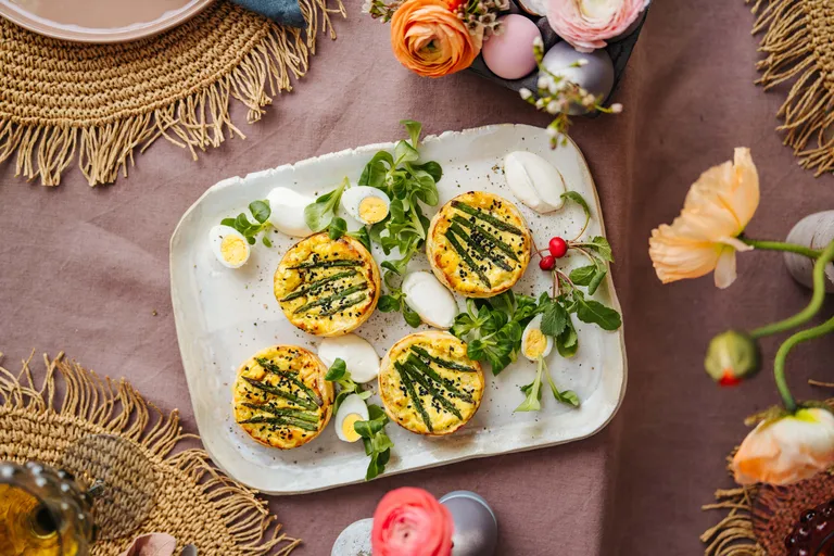Ein weißer Teller mit Mini-Quiches, garniert mit Spargel, umgeben von Blattgemüse, Wachteleiern und Blumen auf einer strukturierten Tischdecke.
