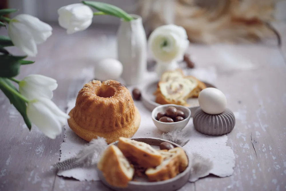 Ein Tisch im Oster-Stil mit Mini-Reindling, Schokoladeneiern, geschnittenem Brot, Keramikeiern und weißen Blumen auf einer rustikalen Unterlage.