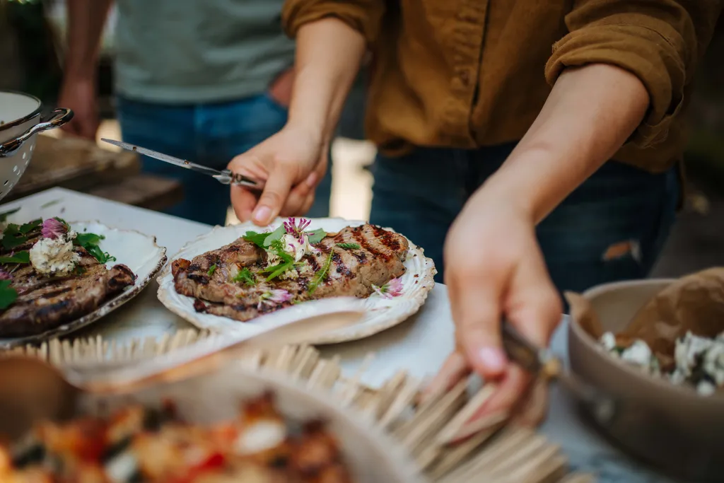 Person serviert frisch gegrilltes Steak mit Kräuterbutter und essbaren Blüten bei einer Grilljause