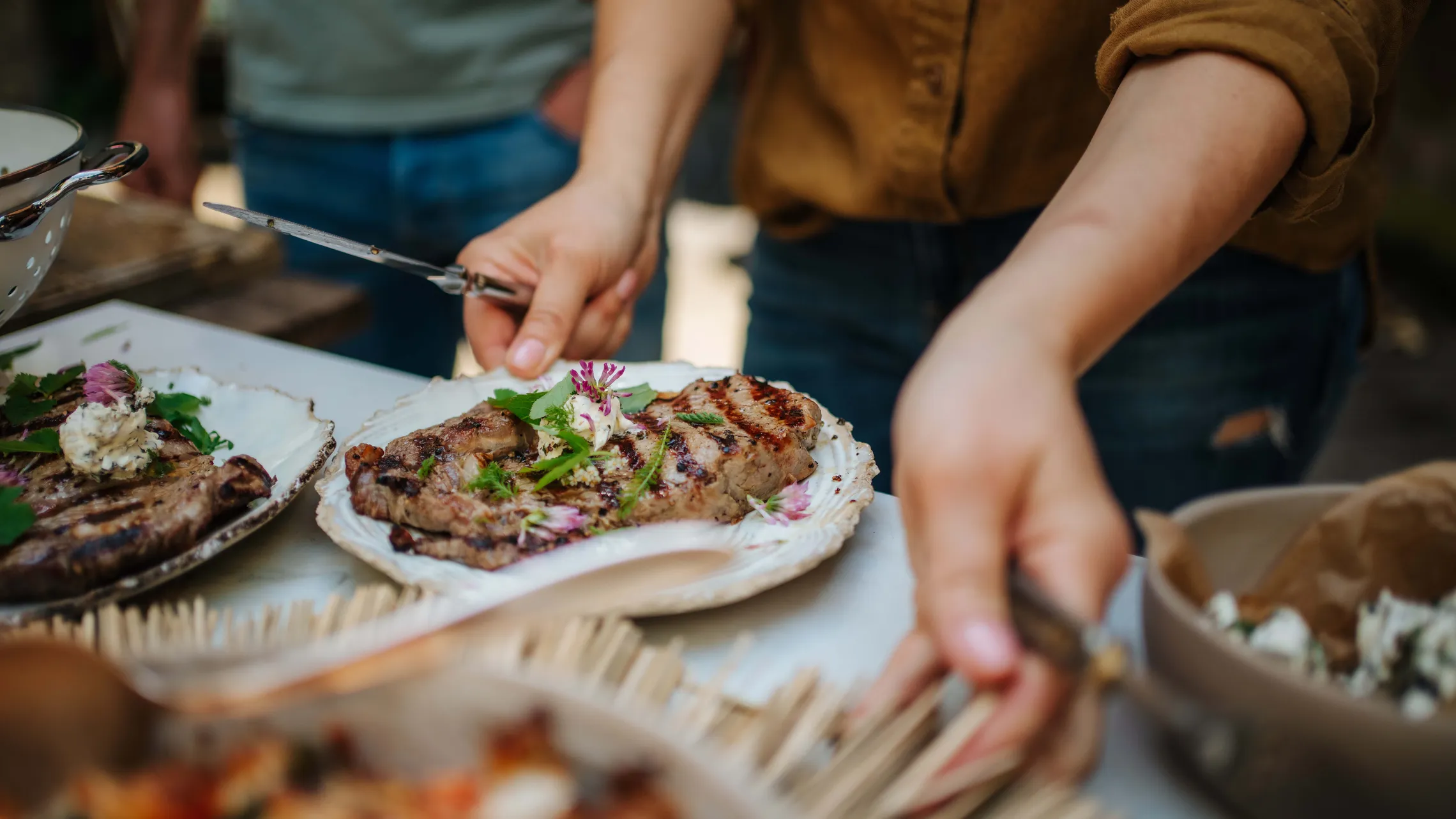 Person serviert frisch gegrilltes Steak mit Kräuterbutter und essbaren Blüten bei einer Grilljause