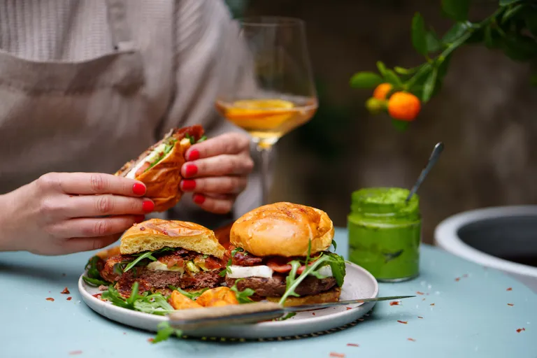 Eine Person, die einen Burger über einem Teller mit Burgern und Salat hält, daneben stehen ein Glas mit grüner Soße und ein Weinglas auf einem Tisch mit dekorativen Orangen.