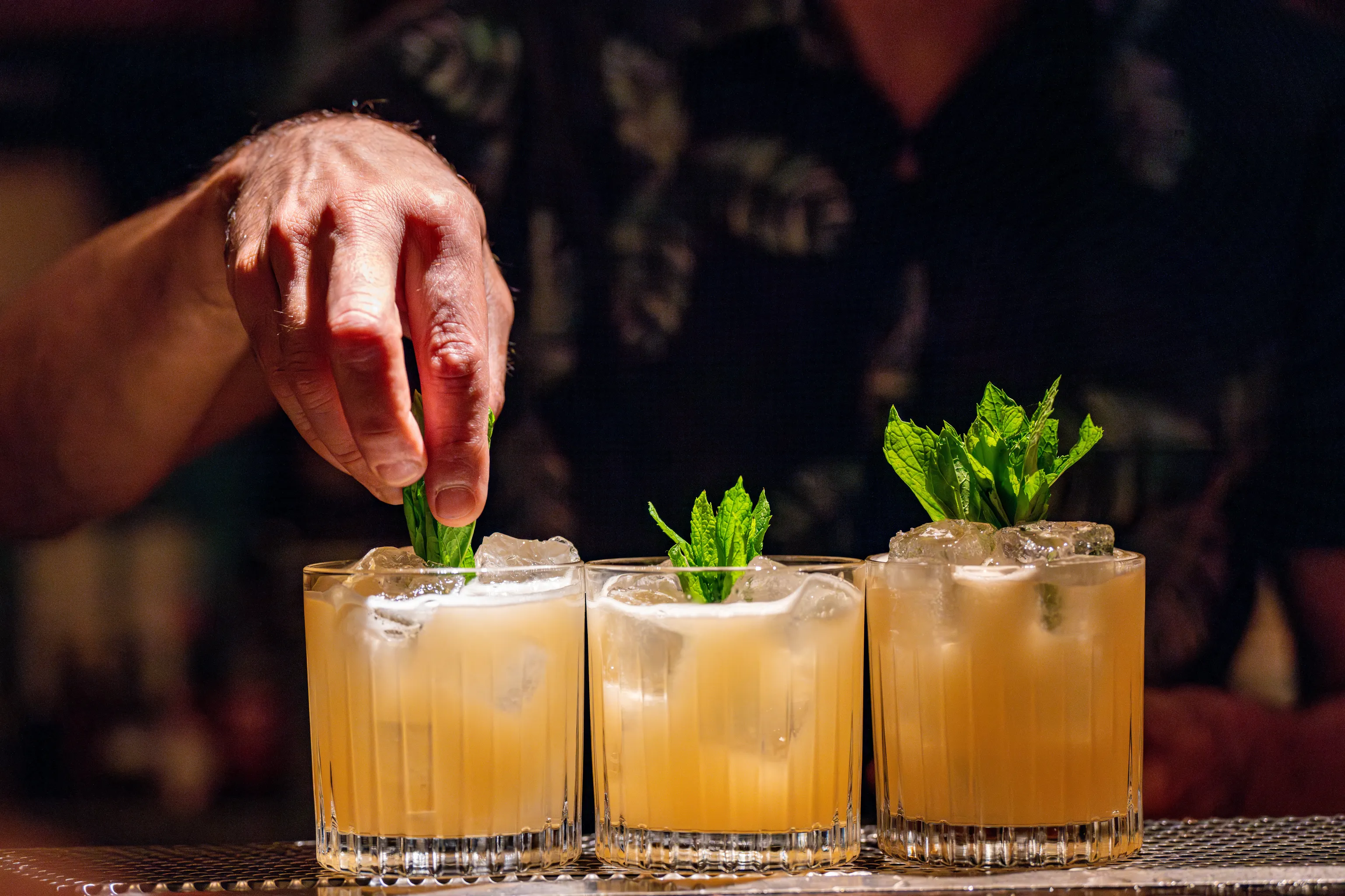 Eine Hand, die einen Cocktail mit Minzblättern garniert; drei Gläser, gefüllt mit einem hellorangen Getränk und Eis, auf einer Bartheke angeordnet.