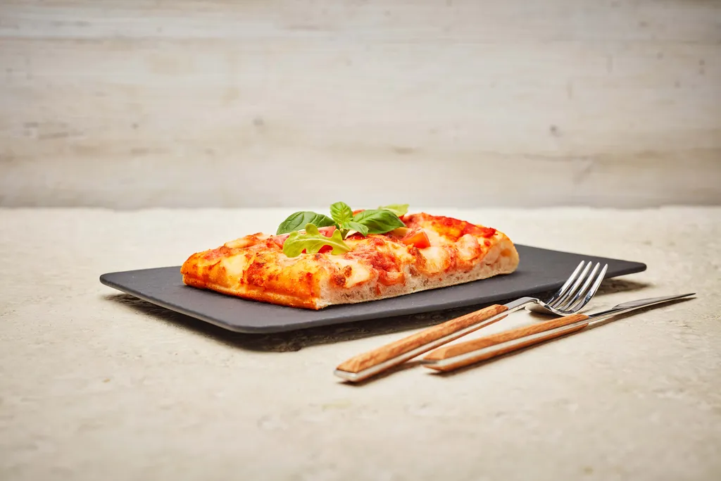 Ein Stück rechteckige Focaccia mit Käse und Basilikum auf einer schwarzen Platte, dazu eine Gabel und ein Messer auf einer leicht strukturierten Oberfläche.