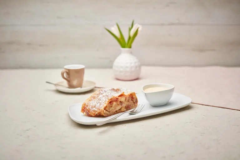 Ein Stück Apfelstrudel mit Puderzucker auf einem Teller, dazu eine kleine Schale mit Soße, eine Gabel, eine Kaffeetasse und eine Vase mit Tulpen.