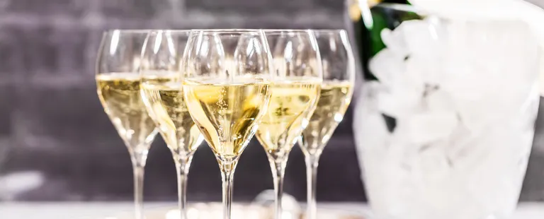 Fünf Champagnergläser, gefüllt mit goldfarbener, sprudelnder Flüssigkeit, und im unscharfen Hintergrund ein Eiskübel, in dem eine Flasche steht.