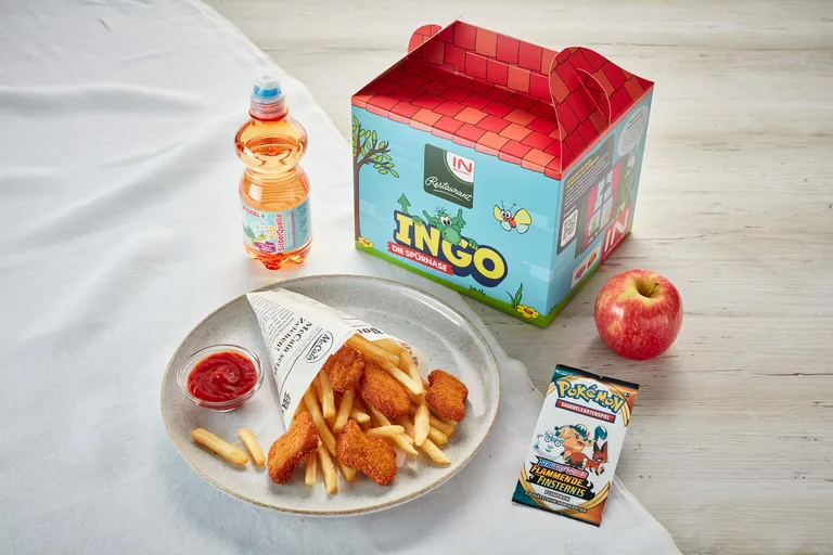 Ein Kindermenü mit Nuggets, Pommes frites, Ketchup, Apfelsaft, einem Apfel, einem Pokémon-Kartenpaket und einer bunten Schachtel auf einem Tisch.
