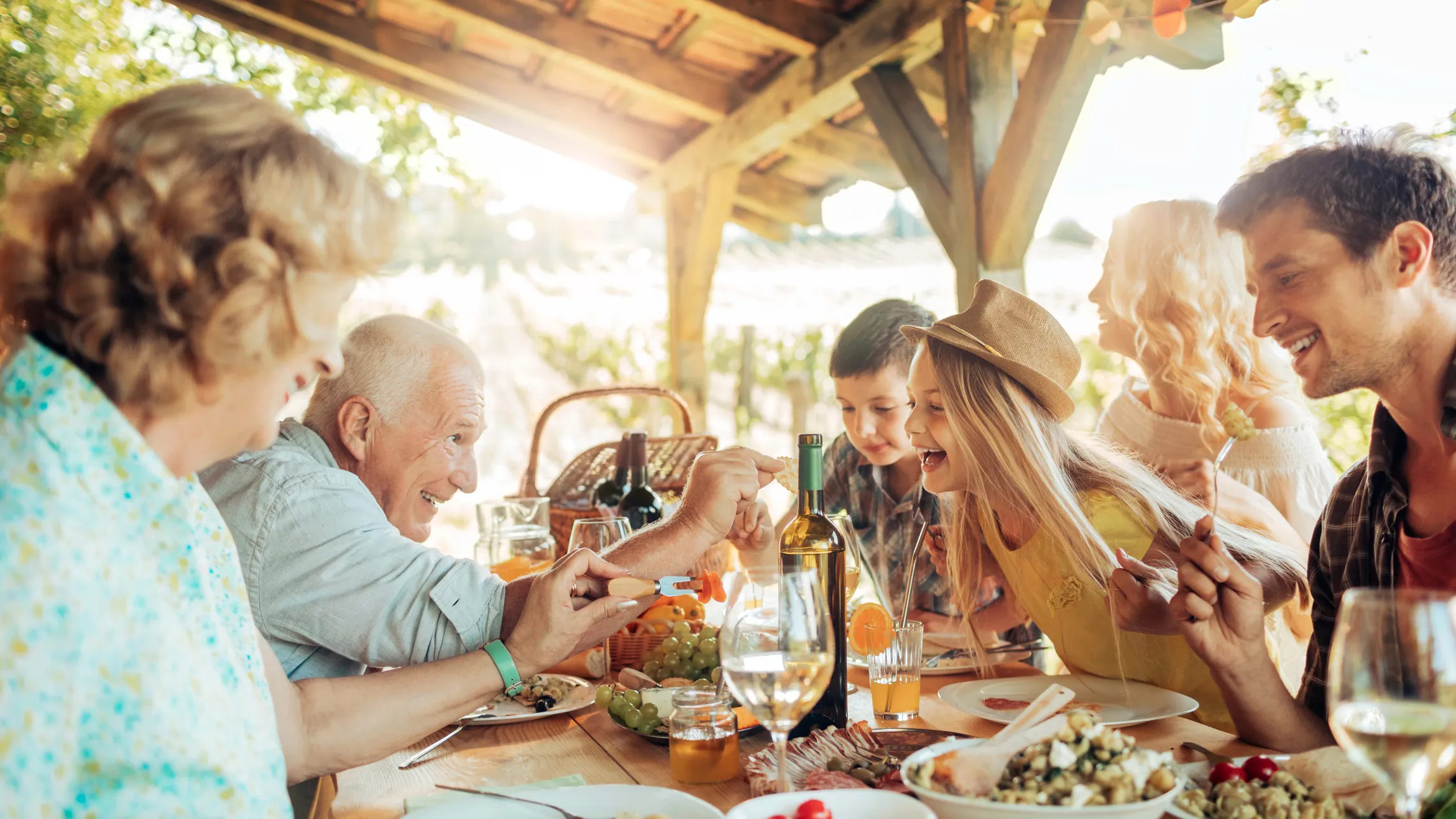 Eine Familie genießt ein fröhliches Essen im Freien unter einem rustikalen Holzpavillon, umgeben von üppigem Grün und einer Vielzahl von Gerichten auf dem Tisch.