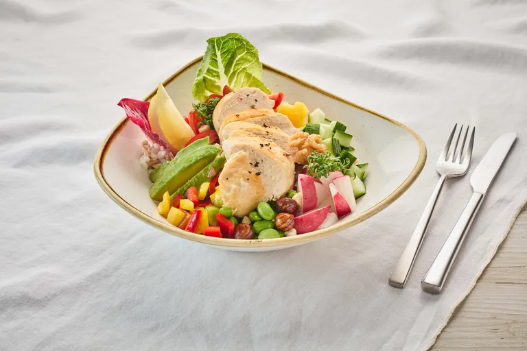 Ein Salat mit Hähnchenstreifen, Avocado, Paprika, Edamame, Radieschen und Gurken auf einem Teller, dazu eine Gabel und ein Messer auf einer weißen Tischdecke.