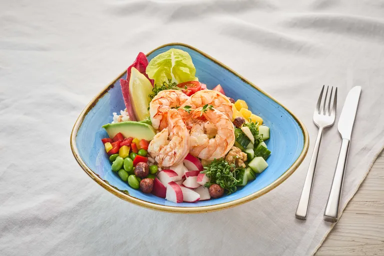 Ein farbenfroher Salat mit Garnelen, Avocado, Radieschen, Edamame, Nüssen und Mango auf einem blauen Teller, der auf einer weißen Tischdecke mit Besteck angerichtet ist.