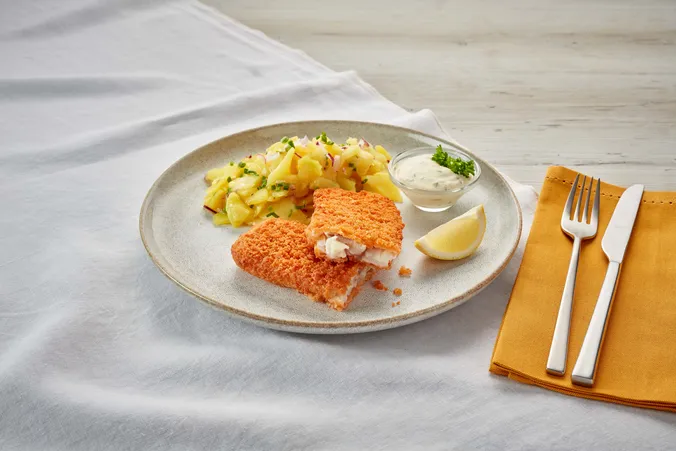 Ein paniertes Fischfilet mit einer Zitronenscheibe, Kartoffelsalat und Tartarsauce auf einem Teller, daneben liegt das Besteck auf einer gelben Serviette.