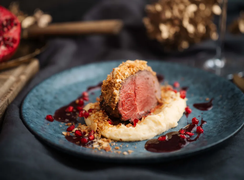 Ein Hirschrückenfilet auf cremigem Püree, garniert mit Granatapfelkernen und Soße, serviert auf einem blauen Teller.