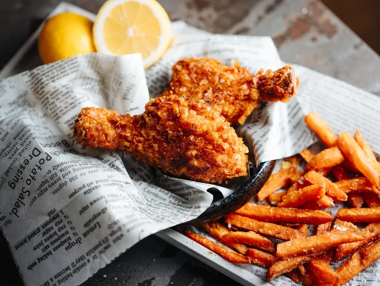 Frittiertes Hähnchen und Süßkartoffel-Pommes frites, serviert auf Zeitungspapier, im Hintergrund eine Zitronenscheibe.