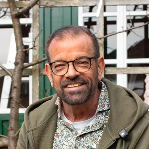 Ein lächelnder Mann in einem Garten, der auf einem hölzernen Hochbeet sitzt, einen Salatkopf in der Hand hält, eine Brille und Gartenhandschuhe trägt, im Hintergrund sind grüne Türen zu sehen.
