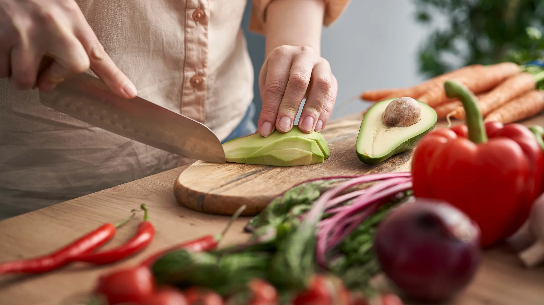 Eine Person schneidet auf einem Holzbrett eine Avocado in Scheiben, umgeben von Karotten und einer roten Paprika; im Vordergrund stehen frische Zutaten und das Kochen.