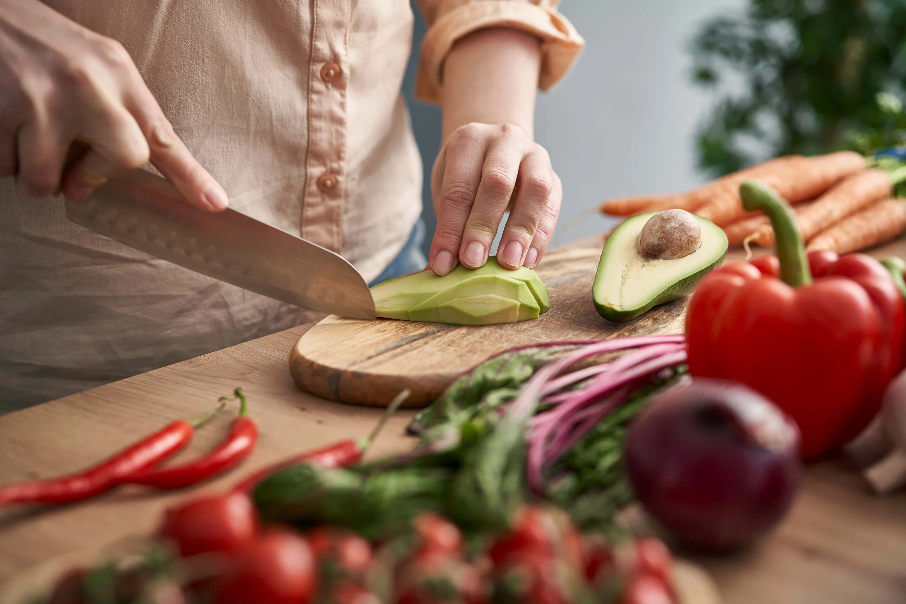 Eine Person schneidet auf einem Holzbrett eine Avocado in Scheiben, umgeben von Karotten und einer roten Paprika; im Vordergrund stehen frische Zutaten und das Kochen.