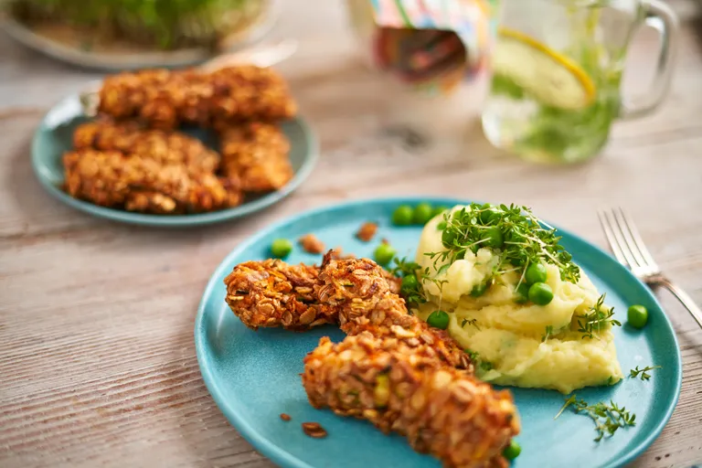 Blauer Teller mit knusprig panierten Gemüsesticks, Kartoffelpüree mit Erbsen und Blattgemüse; im Hintergrund sind ein Getränk und eine Platte zu sehen.
