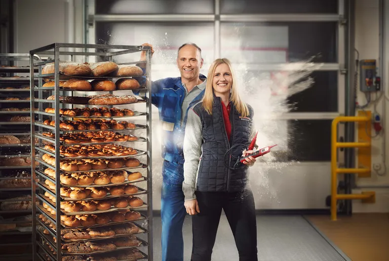 Zwei Mitarbeitende in einer Bäckerei neben einem Wagen mit frisch gebackenen Broten und Gebäck