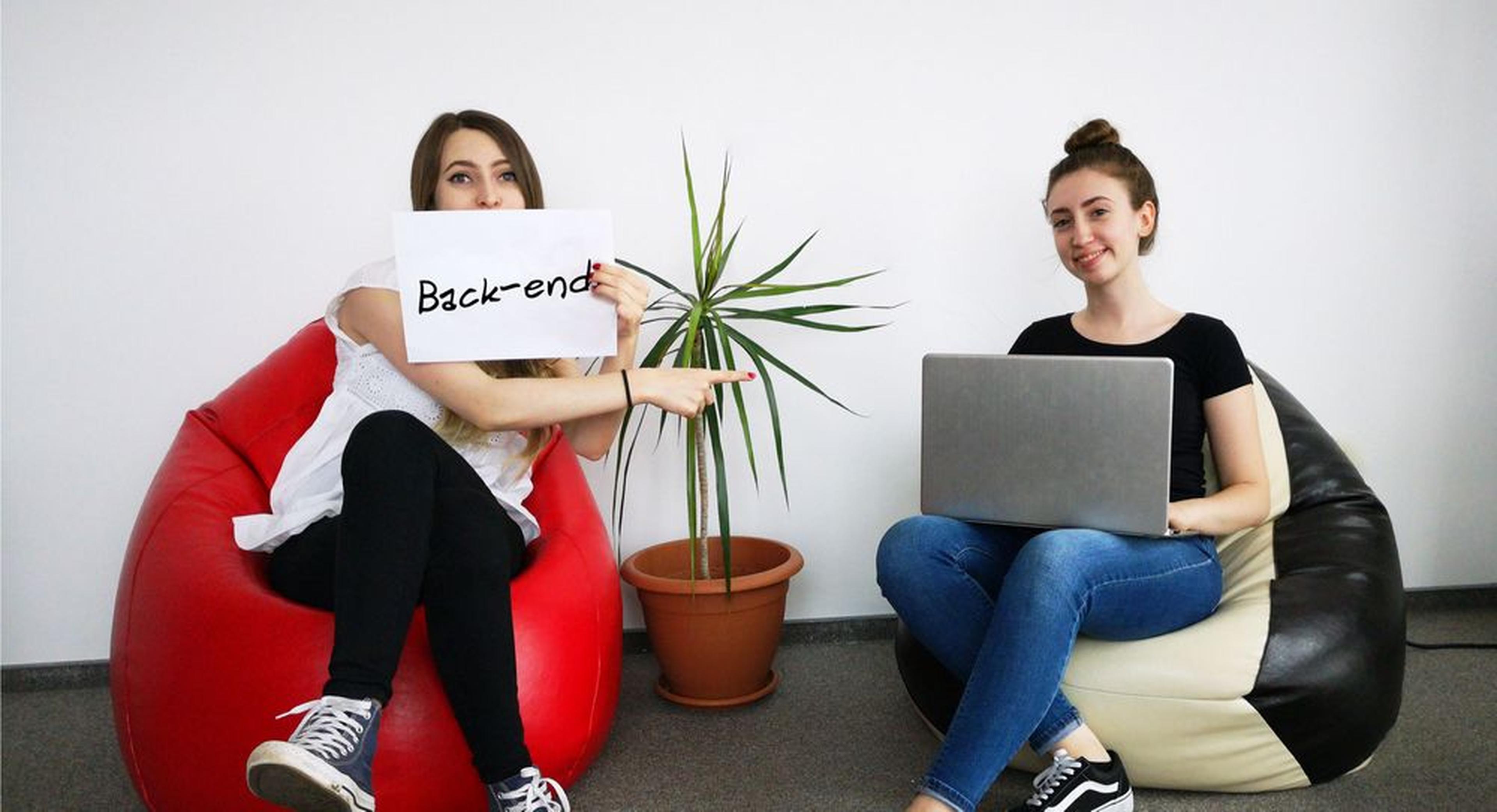 Two women sit on bean bags; one holds a "Back-end" sign, the other uses a laptop. A potted plant is in the background.