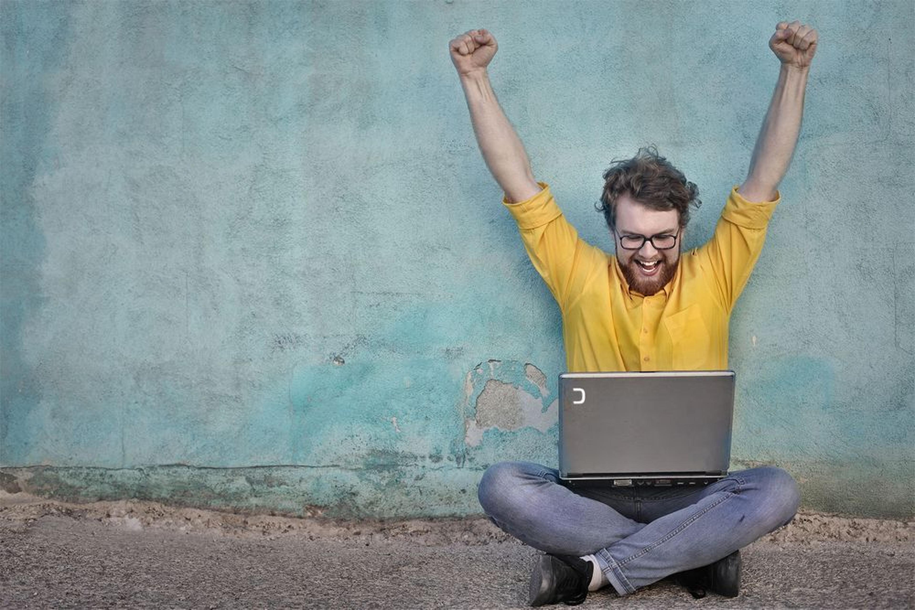 Man in yellow shirt sitting cross-legged, celebrating with arms raised, while using a laptop against a textured blue wall.